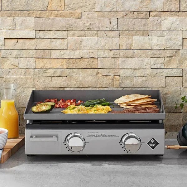 A countertop flat top grill with various foods cooking on it, including eggs, vegetables, and pancakes.