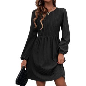 A black, long-sleeve dress with a subtle textured pattern, featuring a round neckline and an A-line silhouette. It is accessorized with a small chain necklace and a quilted black clutch.