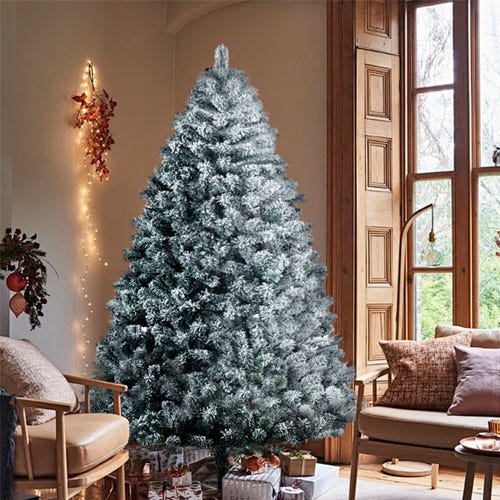 A snow-flocked artificial Christmas tree stands in a cozy living room with wooden furniture and gift boxes underneath. Soft lighting and autumnal decorations add warmth to the room.