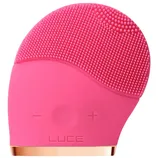 A pink silicone facial cleansing brush with two buttons marked \“-\“ and \“+\“, featuring soft bristles for exfoliating and cleaning the skin. The word \“LUCE\“ is embossed on its front.