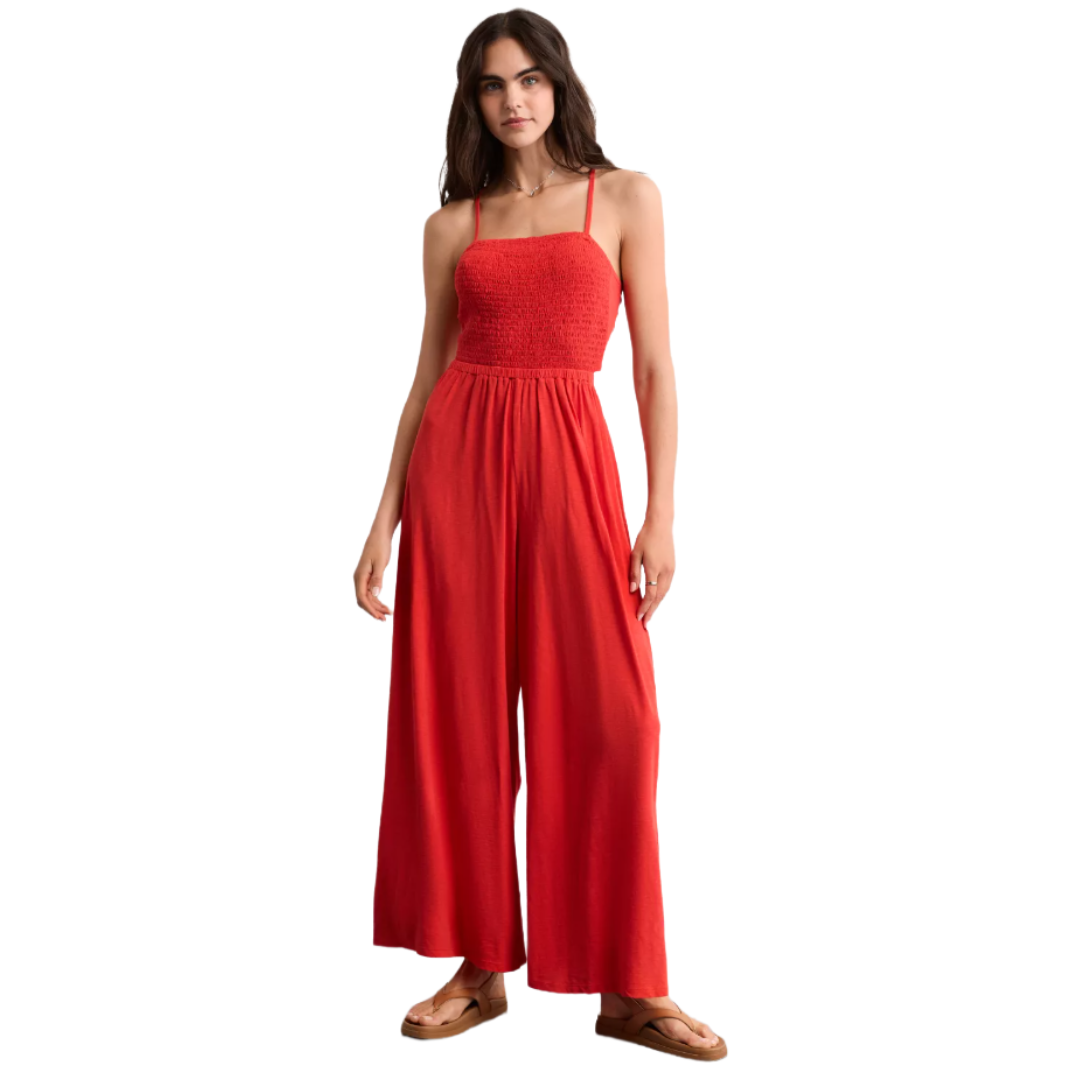 A red sleeveless jumpsuit with a smocked bodice, spaghetti straps, and wide-leg pants, paired with brown sandals.