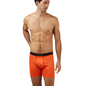 A man models orange boxer briefs with a black waistband featuring the \“32° COOL\“ brand, standing barefoot against a gray background.
