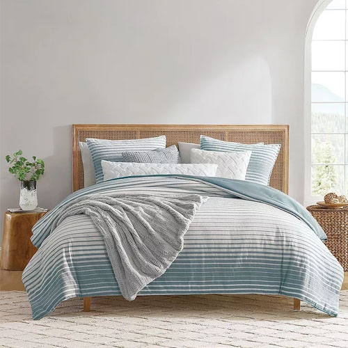 A Koolaburra by UGG Comforter Set featuring a striped pattern in shades of blue and white, with a textured throw blanket draped over, styled on a bed in a bright room.