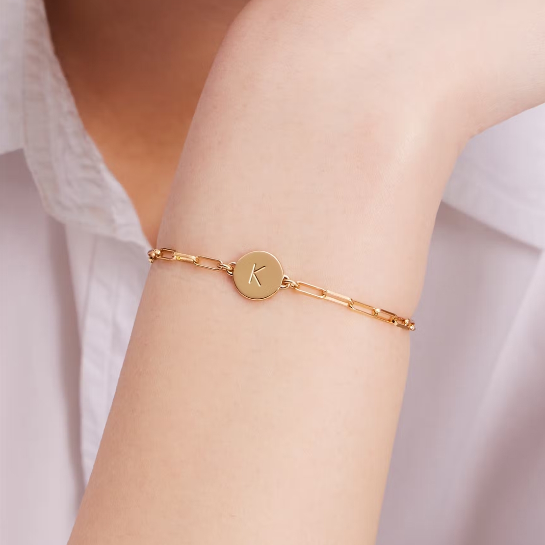 A gold bracelet features a round pendant with the letter \“K,\“ connected by a chain of elongated links, worn on a person's wrist.