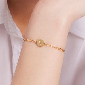 A gold bracelet features a round pendant with the letter \“K,\“ connected by a chain of elongated links, worn on a person's wrist.