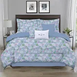 A floral-patterned bedding set with a light blue base includes a comforter, pillow shams, and decorative pillows, arranged on a bed in a modern bedroom setting.
