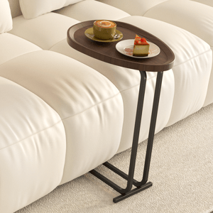 A compact, wooden slide-under side table with a teardrop-shaped top and sleek black metal legs fits snugly against a sofa. Holds a cup of coffee and a small plate with cake, ideal for convenient snacking or working from the couch.