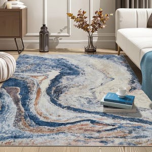 A blue, cream, and brown area rug with a marbled design covers a wooden floor, accompanied by a white sofa and stacked books. A vase with branches is placed nearby for decoration.