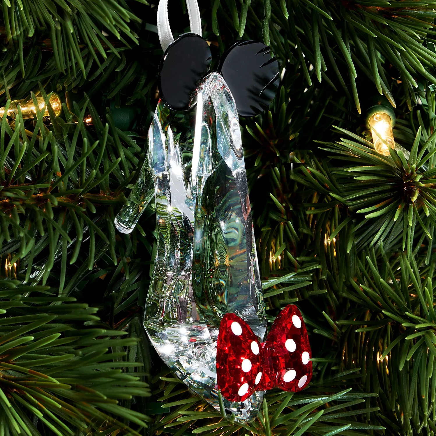 A clear high-heeled shoe ornament with black mouse ears and a red polka-dot bow hangs on a Christmas tree branch.