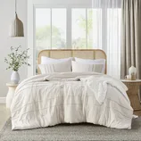 A rattan headboard bed with a white comforter, pillows, a knitted throw, beside a round side table with a vase and decorative items.