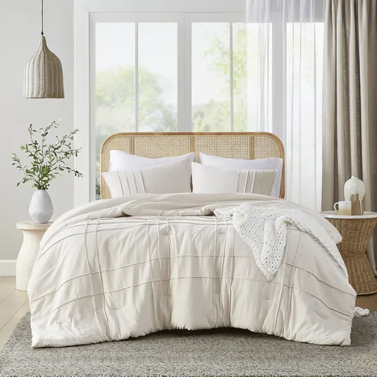A rattan headboard bed with a white comforter, pillows, a knitted throw, beside a round side table with a vase and decorative items.