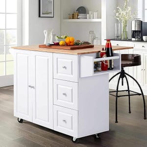 White kitchen island with a wooden countertop, drawers, cabinets, and a side shelf holding condiments. It is equipped with wheels for mobility.