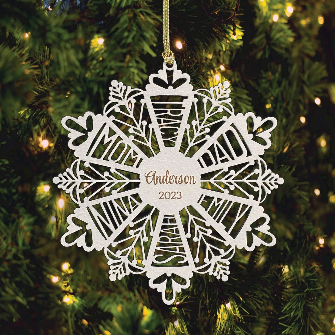 A white snowflake ornament featuring the name \“Anderson\“ and the year \“2023\“ hangs on a Christmas tree with lights.