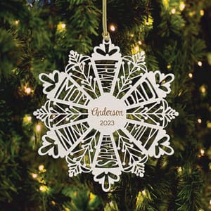 A white snowflake ornament featuring the name \“Anderson\“ and the year \“2023\“ hangs on a Christmas tree with lights.