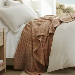 A cozy bedroom scene with a bed featuring white sheets and an earth-toned waffle-textured blanket, a wooden nightstand with books and a lamp, and a large window offering a beach view.