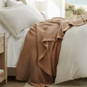 A cozy bedroom scene with a bed featuring white sheets and an earth-toned waffle-textured blanket, a wooden nightstand with books and a lamp, and a large window offering a beach view.