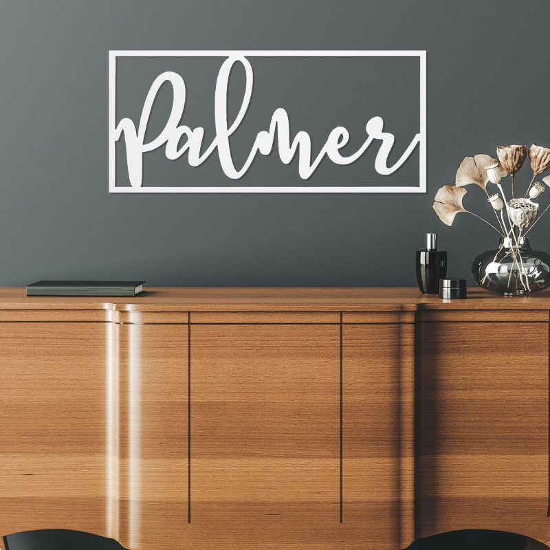 A wooden sideboard is topped with a small stack of books and a decorative vase with dried flowers. The word \“Palmer\“ is prominently displayed on the wall in white cursive within a rectangular frame.