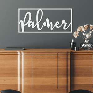 A wooden sideboard is topped with a small stack of books and a decorative vase with dried flowers. The word \“Palmer\“ is prominently displayed on the wall in white cursive within a rectangular frame.
