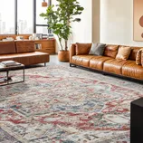 A modern living room features a large patterned rug with blue, red, and beige tones, two brown leather sofas, a tall potted plant, and abstract wall art. A cityscape view is visible through large windows.