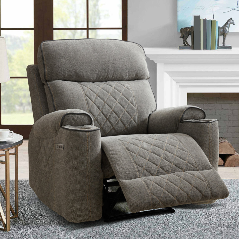 A gray, quilted recliner chair is placed in a living room setting with a side table, a lamp, and decorative items such as books and a sculpture on a mantle.