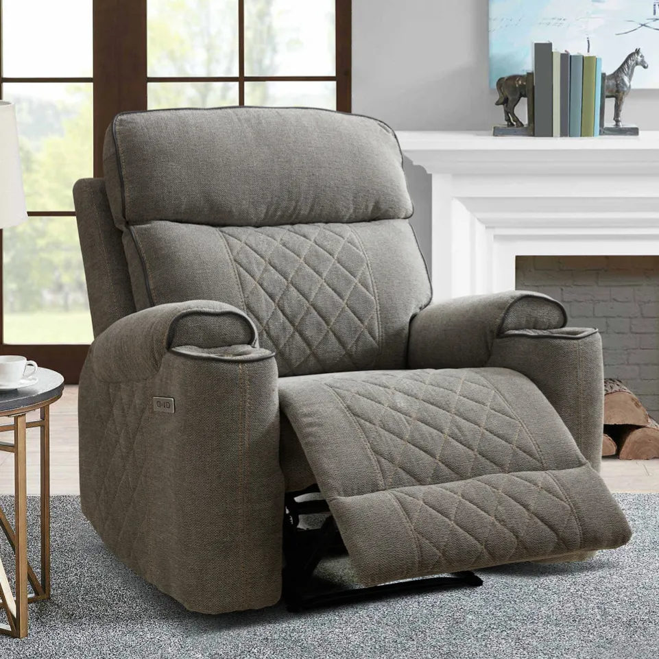 A gray, quilted recliner chair is placed in a living room setting with a side table, a lamp, and decorative items such as books and a sculpture on a mantle.