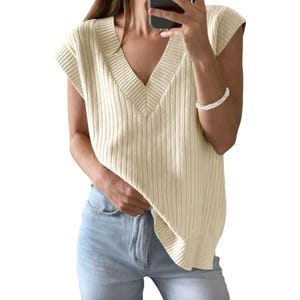 A sleeveless, ribbed knit sweater in cream with a V-neck design is paired with light blue jeans.