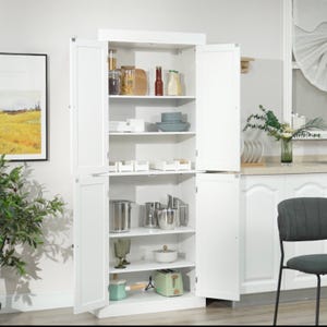 White kitchen cabinet with doors open, displaying dishes, glasses, and cooking pots.