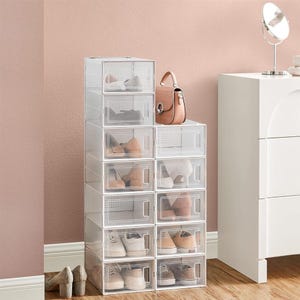 A tower of clear, stackable shoe storage boxes with a small pink handbag on top, placed next to a white cabinet with a mirror.