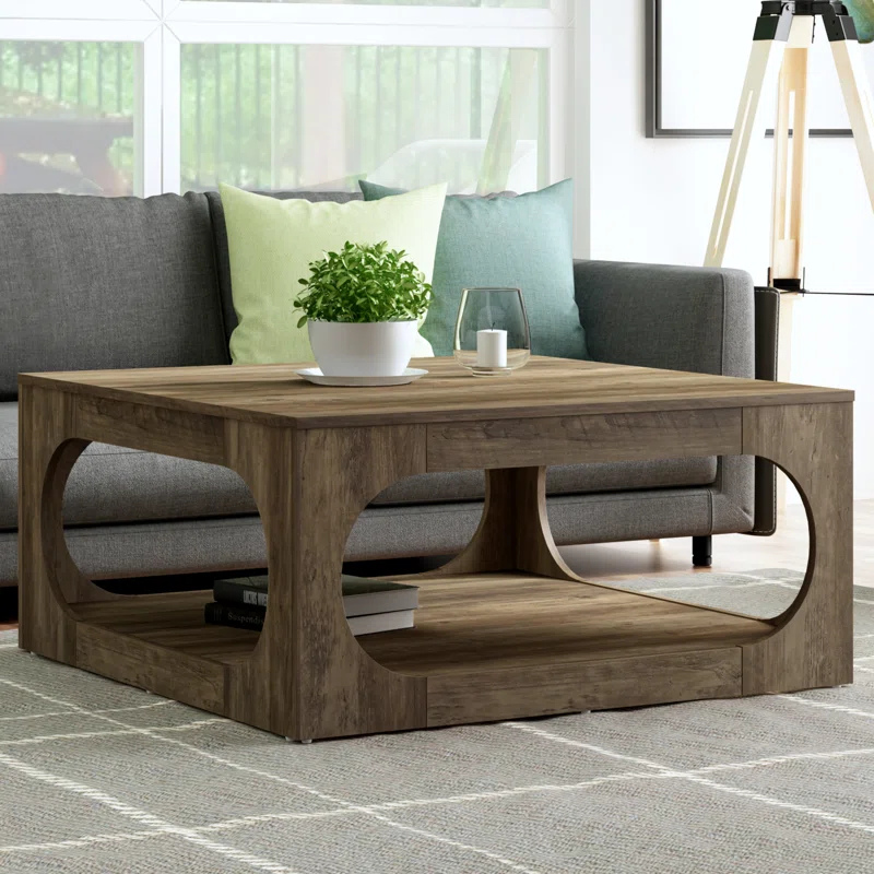 A wooden coffee table with an open geometric design, featuring circular cutouts and a lower shelf. It is placed in front of a gray couch and is adorned with a small plant and a glass candle holder on top.