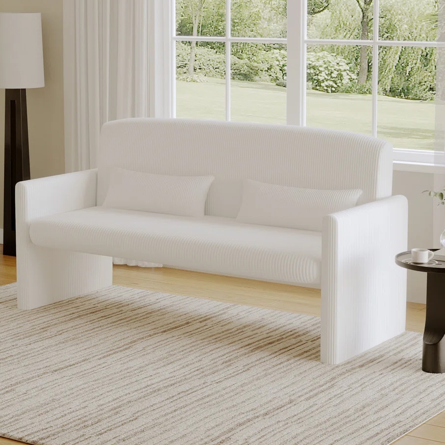 A modern white loveseat with vertical ribbed texture, featuring two matching rectangular throw pillows, set on a light striped rug in a bright room with large windows and natural light.