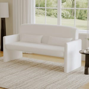 A modern white loveseat with vertical ribbed texture, featuring two matching rectangular throw pillows, set on a light striped rug in a bright room with large windows and natural light.