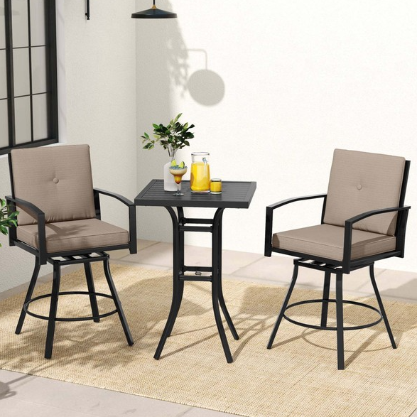 A patio bar set featuring a high square table and two swivel chairs with beige cushions, all in a black metal frame. The table holds a vase of green leaves and a tray with a pitcher and glasses. The set is placed on a light woven rug.