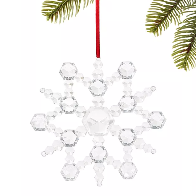 A clear crystal snowflake ornament with distinct facets hangs from a red ribbon, accompanied by green pine foliage in the background.