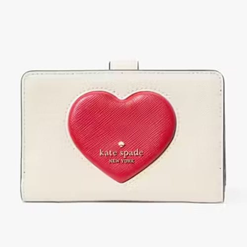 A compact cream-colored wallet featuring a prominent red puffy heart design, emblazoned with the \“kate spade new york\“ logo on the front.