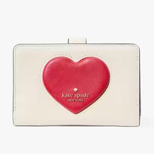 A compact cream-colored wallet featuring a prominent red puffy heart design, emblazoned with the \“kate spade new york\“ logo on the front.