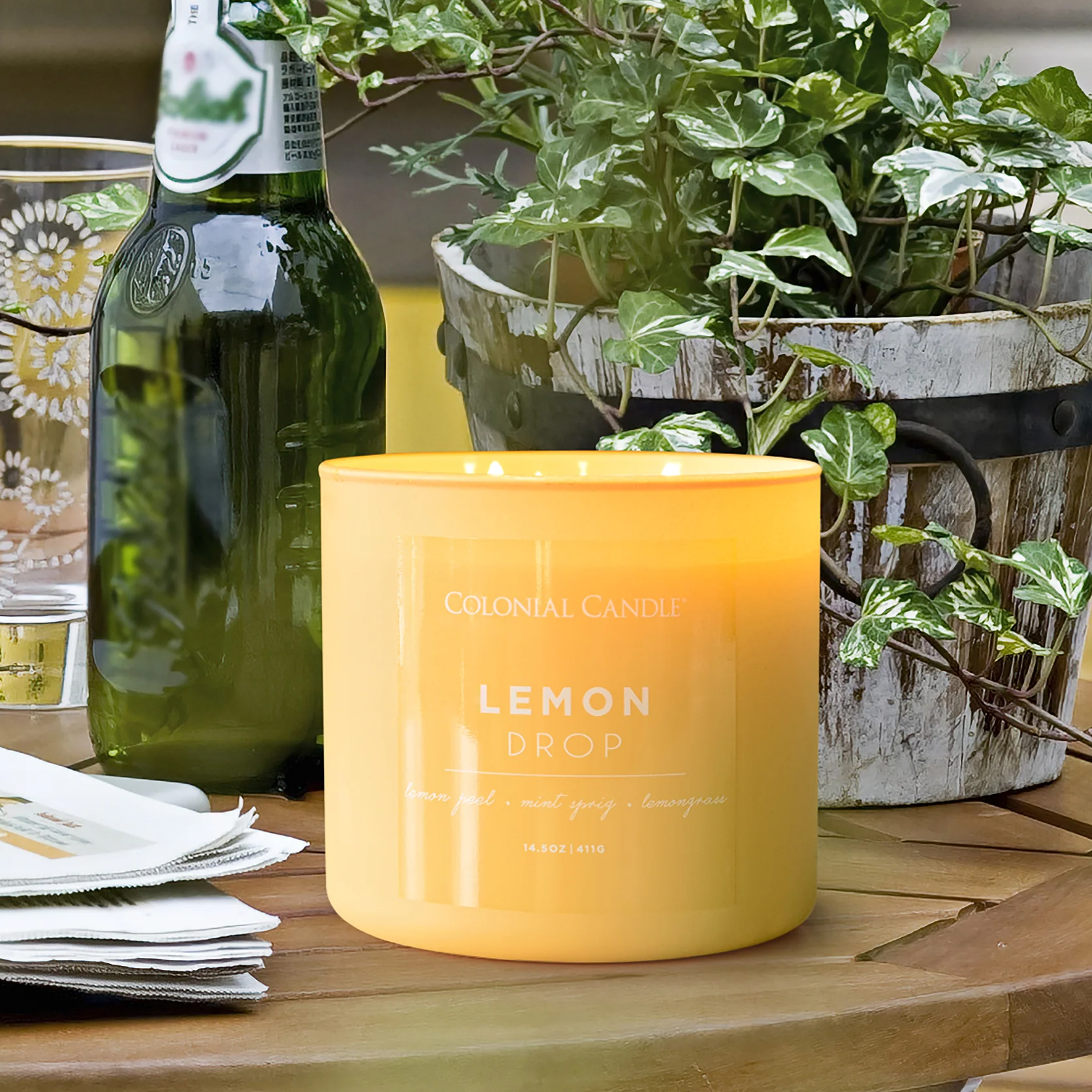 A Colonial Candle labeled \“Lemon Drop\“ with scents of lemon peel, mint sprig, and lemongrass is on a table next to a bottle, a patterned glass, and a plant in a wooden container.