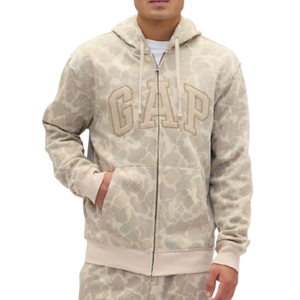 A person is wearing a beige camouflage hoodie with \“GAP\“ embroidered on the chest. The hoodie features a front zipper, drawstrings, and front pockets.