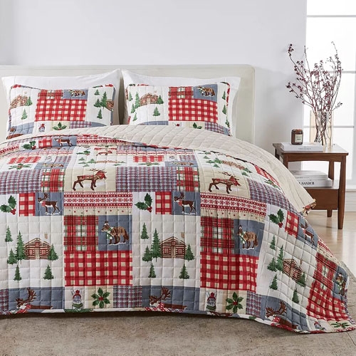 A bedding set featuring a patchwork quilt with red and blue plaid patterns, and nature-themed designs like trees, moose, and cabins, accompanied by matching pillow shams on a neatly made bed in a cozy bedroom setting.