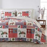 A bedding set featuring a patchwork quilt with red and blue plaid patterns, and nature-themed designs like trees, moose, and cabins, accompanied by matching pillow shams on a neatly made bed in a cozy bedroom setting.