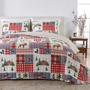 A bedding set featuring a patchwork quilt with red and blue plaid patterns, and nature-themed designs like trees, moose, and cabins, accompanied by matching pillow shams on a neatly made bed in a cozy bedroom setting.