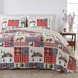 A bedding set featuring a patchwork quilt with red and blue plaid patterns, and nature-themed designs like trees, moose, and cabins, accompanied by matching pillow shams on a neatly made bed in a cozy bedroom setting.