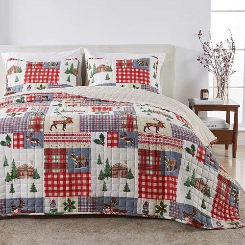 A bedding set featuring a patchwork quilt with red and blue plaid patterns, and nature-themed designs like trees, moose, and cabins, accompanied by matching pillow shams on a neatly made bed in a cozy bedroom setting.