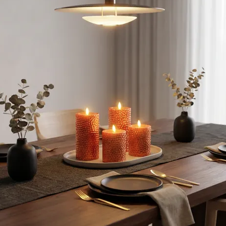 A dining table setup with textured orange candles on a tray, flanked by black vases with dried foliage, under a modern lamp.