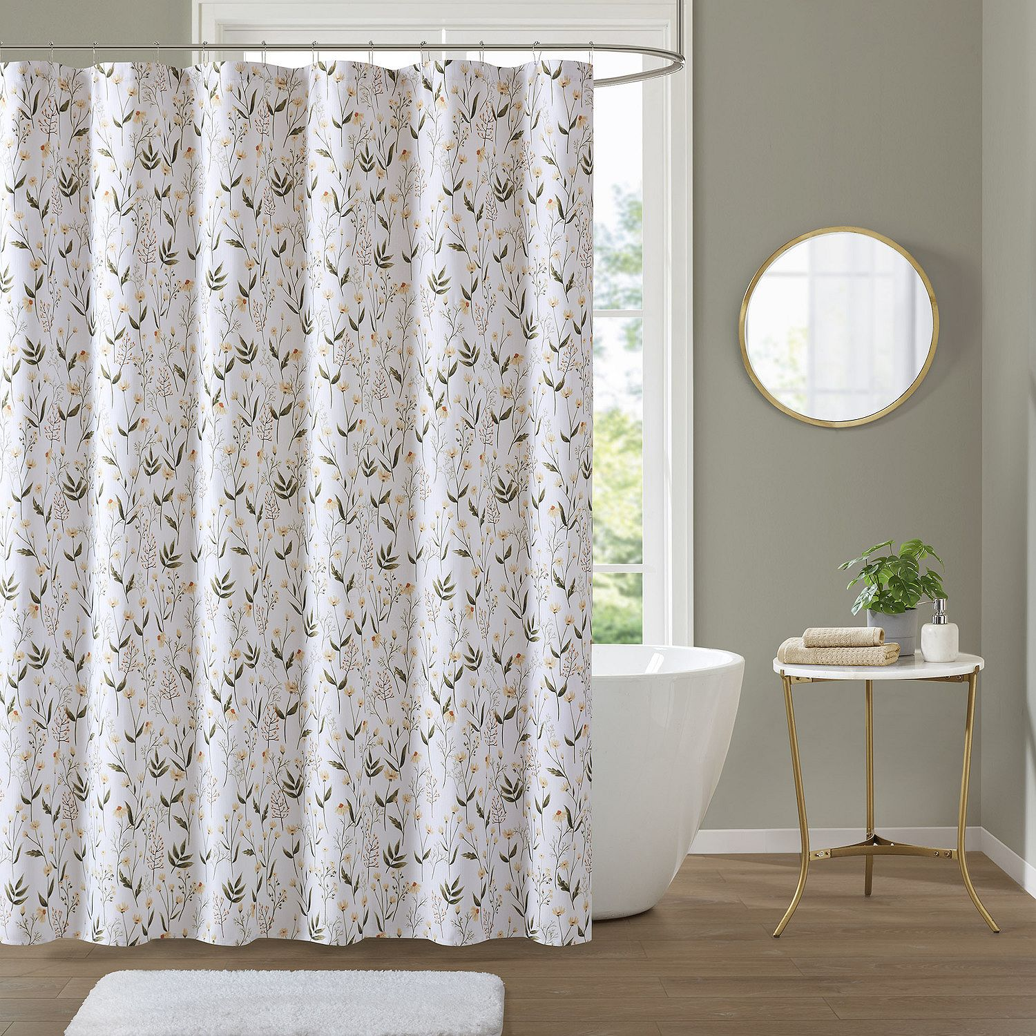 Floral shower curtain with a subtle pattern, alongside a white bathtub, round mirror, small table with plant, and towels, on a light wooden floor.