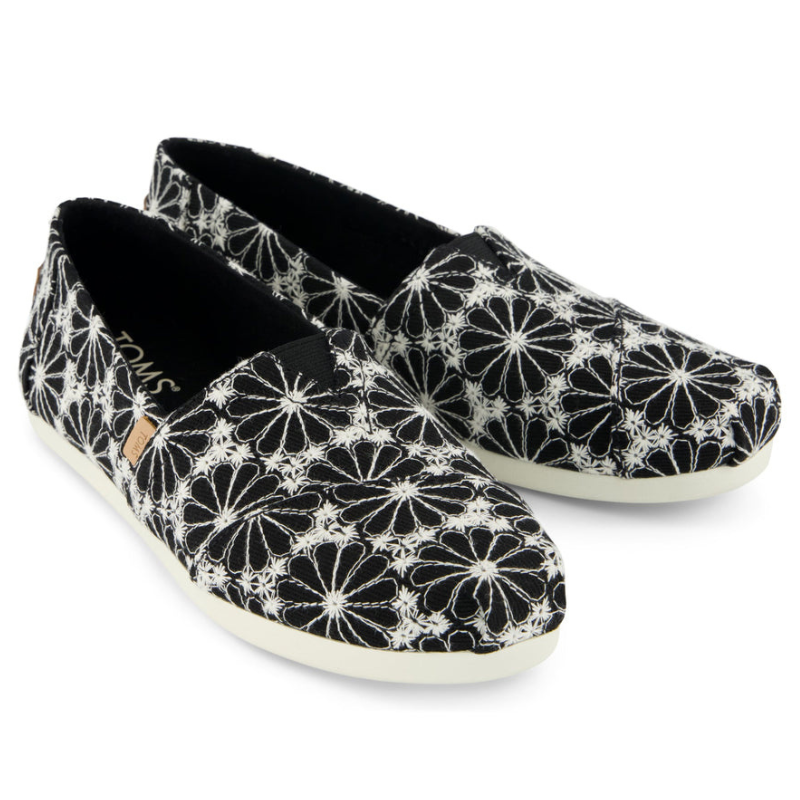 Black slip-on shoes with a white floral pattern, featuring a low-profile sole and fabric upper.