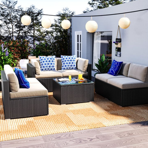 Outdoor patio set with beige cushions and blue patterned throw pillows includes a wicker sofa, glass-top coffee table, and adjacent chair. String lights and greenery add ambiance, set on a woven rug over wooden decking.