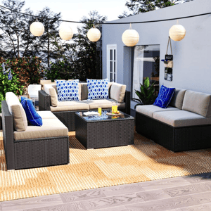 Outdoor patio set with beige cushions and blue patterned throw pillows includes a wicker sofa, glass-top coffee table, and adjacent chair. String lights and greenery add ambiance, set on a woven rug over wooden decking.