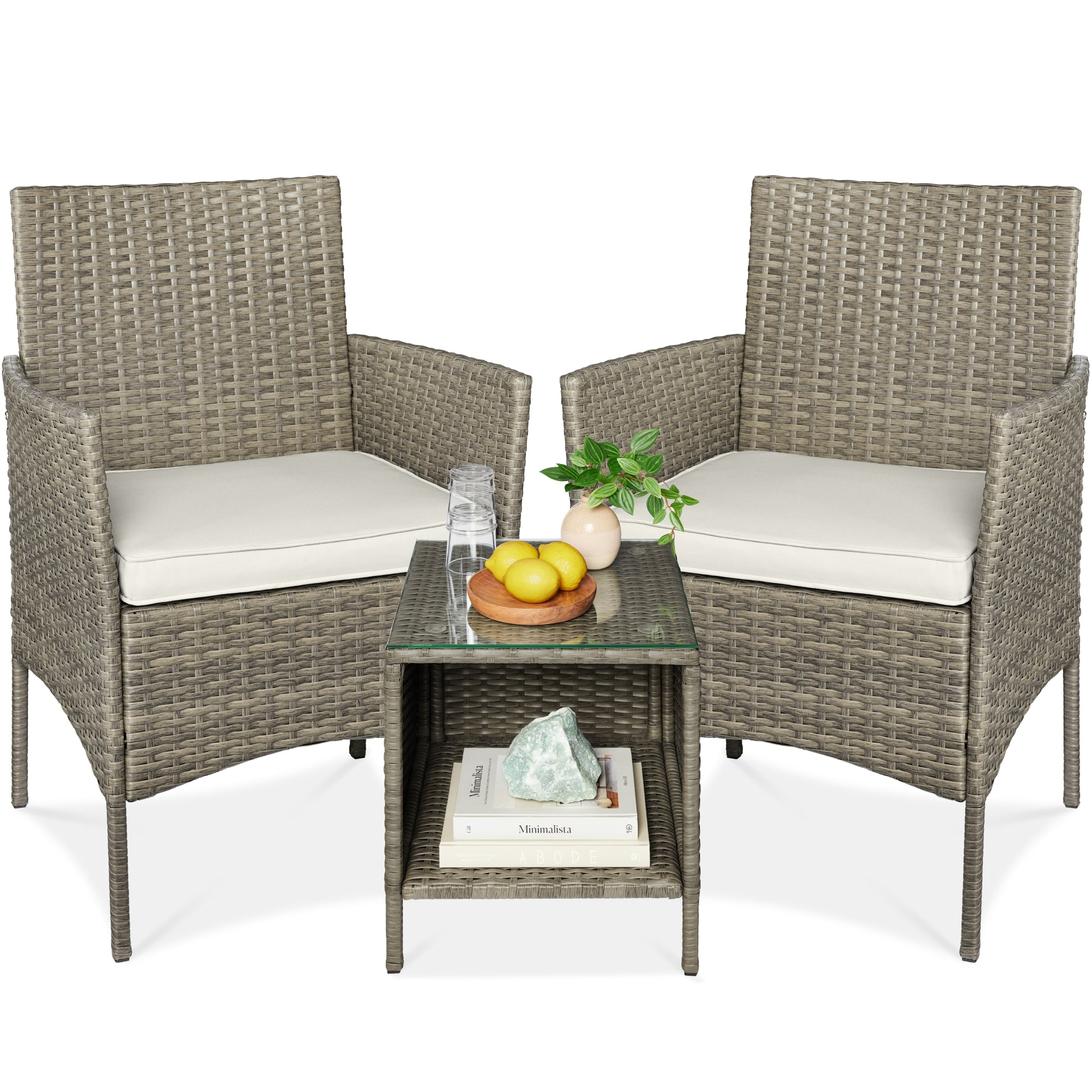 Two wicker chairs with beige cushions are placed alongside a wicker side table featuring a glass top. The table holds a plate of lemons and a small plant, while a shelf below contains books and a decorative rock.