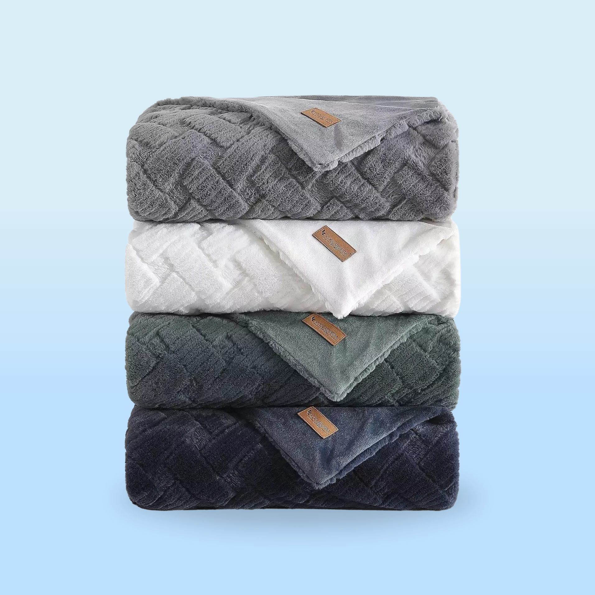 A stack of four fluffy blankets in gray, white, green, and blue hues, each with a textured pattern and a small leather tag.