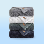 A stack of four fluffy blankets in gray, white, green, and blue hues, each with a textured pattern and a small leather tag.